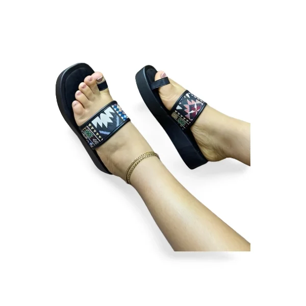 Pieds chaussés de sandales femme à entre-doigt noires, dotées d'une large bride avec motifs ethniques colorés (bleu, marron, blanc, orange) et une semelle épaisse. Une chevillère dorée est visible sur l'une des chevilles, couleur noir